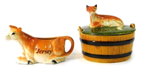 Two Jersey cow ceramics, comprising a seated Jersey cow milk jug, 14cm wide, and a Jersey cow barrel form butter dish, 12cm high. (2)