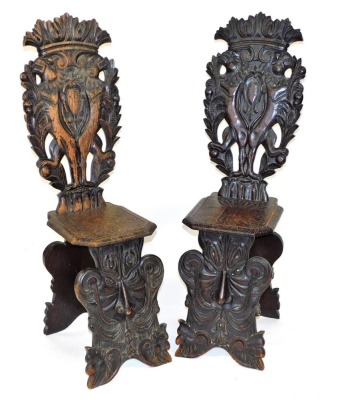 A pair of early 19thC Italian oak Sgabello hall chairs, with foliate carved crest rails, the back carved with a pair of rampant lions holding branches of pomegranates, flanking a central shield, with an engraved solid seat, raised on a grotesque mask base