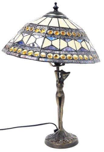 A Tiffany style table lamp, with a geometric panelled and jewel shade, raised on a stem fronted by a nude figure of a lady, above a circular base, 45cm high.