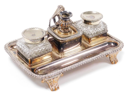 An early 19thC silver plated desk stand, with two pen recesses, pair of cut glass inkwells, with hinged lids, flanking a central container surmounted with a chamber stick and snuffer, raised on scroll feet, 22cm wide.