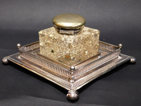 An Edward VII silver plated and cut glass ink stand, by Mappin & Webb, the diamond shaped inkwell, with plain circular hinged lid, on a galleried stand with bright cut arrow outline on compressed orb feet, 9cm high.