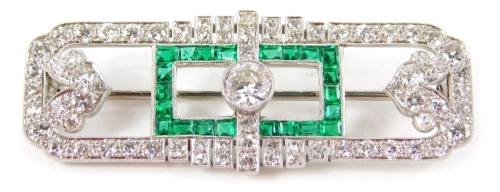 An Art Deco diamond and emerald bar brooch, centred by a central diamond, surrounded by 22 baguette cut emeralds, with a spade and scroll shaped arrangement of further diamonds to each side and a further diamond border, with a plain pin back, unmarked, 5c